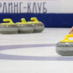 Женская рука запускает камень для кёрлинга на льду Шуваловского льда, на фоне инвентарь и надпись "Кёрлинг-клуб"